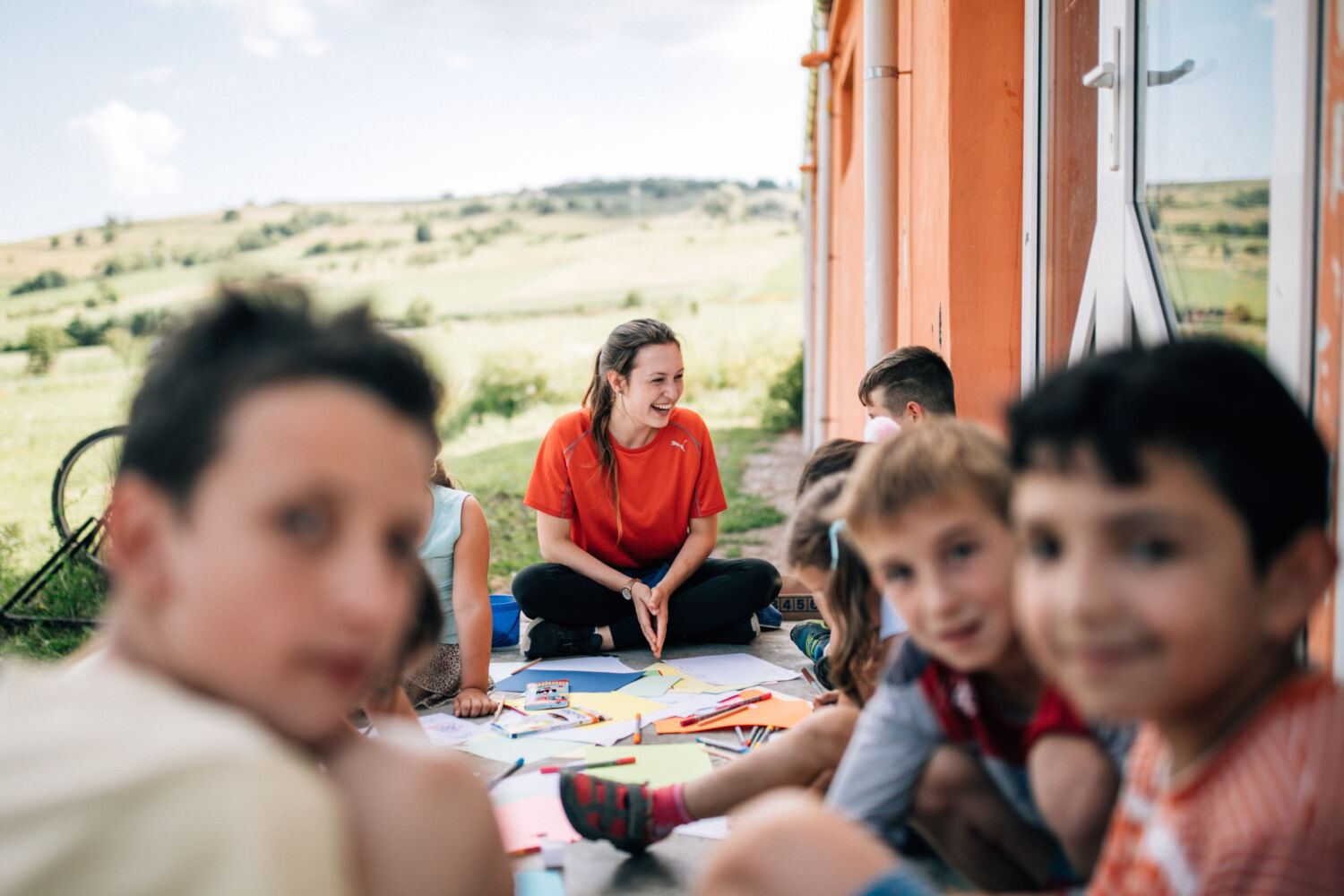 A volunteer sitting with children over craft materials under an open canopy, meadow behind