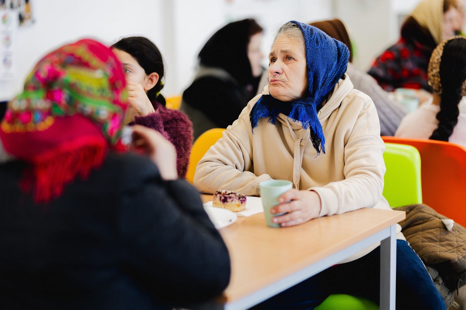An elderly woman in a blue headscarf with a cup of tea and a slice of cake